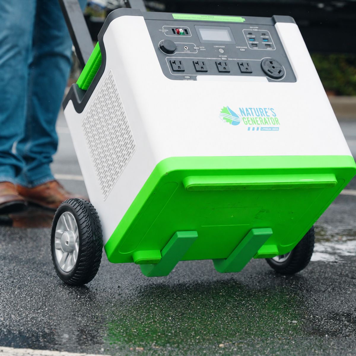 Scene - A Nature's Generator portable power station with green and white panels, wheeled design, and a control panel, being pushed on wet pavement by a person whose legs are partially visible.