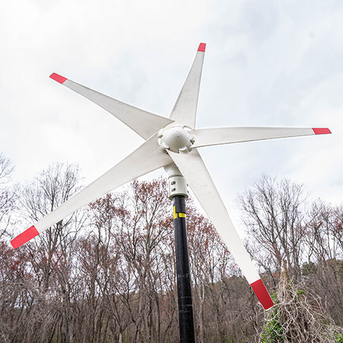 Nature's Generator 150W Wind Turbine - Scene - Wind turbine with red blades against a backdrop of leafless trees and a cloudy sky. 9